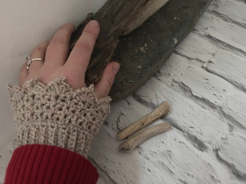 May include: A close-up of a person's hand wearing a beige crocheted wrist warmer with a scalloped edge. The hand is resting on a piece of driftwood.
