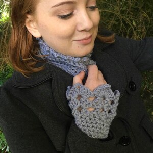 May include: A woman wearing a grey crocheted cowl neck scarf and matching fingerless gloves. The scarf and gloves are made with a delicate open stitch pattern.