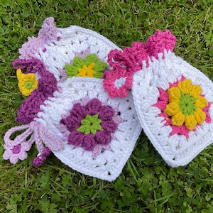 Può includere: Due sacchetti a forma di fiore all'uncinetto con filato bianco, rosa, viola, giallo e verde. I sacchetti sono sdraiati su un letto di erba verde.