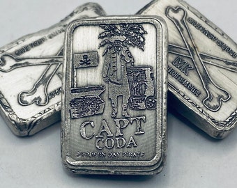 May include: Three silver bars with pirate-themed designs. The central bar features a detailed engraving of a pirate, a treasure chest, and the text "CAPT CODA MODERN DAY PIRATE." The other bars display crossed bones and the text "ONE TROY OUNCE."