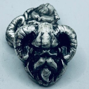 May include: A detailed, silver-toned metal sculpture of a ram's head with prominent horns and a textured surface. The sculpture has a weathered appearance, with intricate detailing on the face and horns. The ram's head is the focal point.