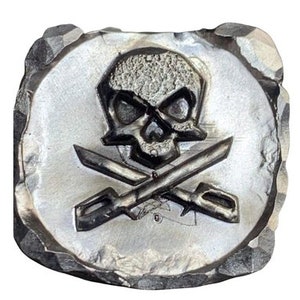 May include: A silver-colored, square-shaped emblem with a skull and crossbones design. The skull has detailed eye sockets and a textured surface. Two crossed swords are beneath the skull. The emblem has a rough, textured border.