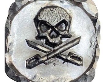 Könnte beinhalten: Ein silberfarbenes, quadratisches Emblem mit einem Totenkopf- und gekreuzten Knochen-Design. Der Schädel hat detaillierte Augenhöhlen und eine strukturierte Oberfläche. Zwei gekreuzte Schwerter befinden sich unter dem Schädel. Das Emblem hat einen rauen, strukturierten Rand.