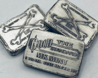 May include: Several silver bars are shown, each stamped with text including "ONE TROY OUNCE" and "MK .999 FINE SILVER". One bar reads "GOLD THE STANDARD MK MINT 1 TR OZ. FINE SILVER 999". The bars feature a skull and crossbones motif.
