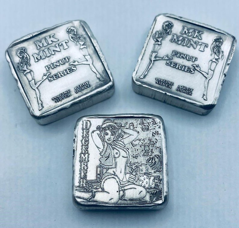 K&ouml;nnte beinhalten: Drei Silberbarren mit eingravierten Pin-up-Designs. Zwei Barren zeigen den Text "MK MINT PINUP SERIES" und zwei Pin-up-Figuren. Der dritte Barren hat ein Dezember-Pin-up-Design mit dem Wort "Dezember" vertikal an der Seite.