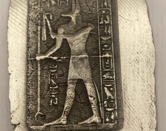 May include: A silver-colored rectangular bar with an embossed image of an Egyptian figure holding a staff. The figure is in a rectangular frame with hieroglyphs. The bar has a rough, textured surface.
