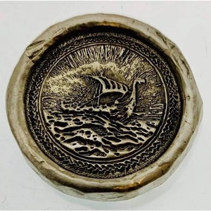 May include: A silver-toned wax seal with a detailed engraving of a Viking ship sailing on choppy waters. The circular seal has a textured border and a weathered appearance, suggesting age and history.