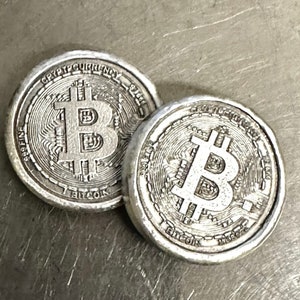Könnte beinhalten: Zwei silberne Bitcoin-Münzen mit detaillierten Gravuren. Die Münzen zeigen das Bitcoin-Symbol und die Wörter "CRYPTOCURRENCY" und "BITCOIN" an den Rändern. Die Münzen liegen auf einer strukturierten Oberfläche.