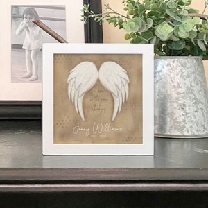 May include: White framed picture with a brown background and white angel wings. The text reads "I am with you always" and "Jenny Williams 1922-2020".