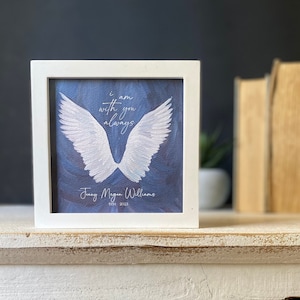 May include: White framed picture with a blue background and white angel wings. The text reads "I am with you always" and "Jenny Megan Williams 1934 - 2023".