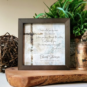 May include: Square framed artwork with a cross design and inspirational text. The dark brown frame surrounds a print with a textured background and the words "I have called you by name..." and the name "Edward Stevenson".