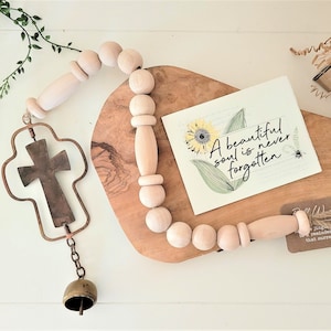 May include: A wooden bead garland with a cross charm and a bell hanging from it. The garland is on a wooden cutting board with a card that reads "A beautiful soul is never forgotten".