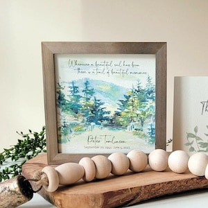 May include: A framed watercolour painting of a forest scene with the quote "Wherever a beautiful soul has been, there is a trail of beautiful memories."  The frame is brown and the painting is on a white background. The text "Peter Tomlinsom, September 20, 1945 - June 5, 2022" is printed below the painting. A small card with the text "Thinking of you" is also visible in the image.