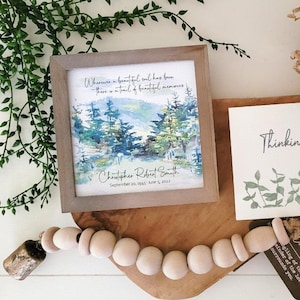 May include: A wooden framed print with a watercolour painting of a forest scene. The print has the text "Wherever a beautiful soul has been, there is a trail of beautiful memories." and "Christopher Robert Smith September 20, 1945 - June 5, 2023". A white card with the text "Thinking of you" and a green floral design is next to the print. A wooden bead garland with a small bell is on a wooden chopping board.