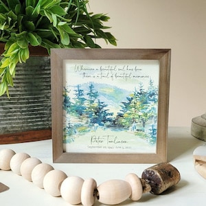May include: A framed watercolour painting of a forest scene with a quote about beautiful souls leaving behind beautiful memories. The quote reads "Wherever a beautiful soul has been, there is a trail of beautiful memories." The frame is brown wood and the painting is signed "Peter Tomlinson" with the date "September 18th 1941 - June 5, 2023".