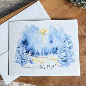 May include: A watercolor Christmas card with a winter forest scene in blue and white, featuring a bright yellow star and the text "O Holy Night." The card is accompanied by a white envelope, set on a wooden surface.