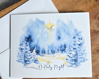 May include: A watercolor Christmas card with a winter forest scene in blue and white, featuring a bright yellow star and the text "O Holy Night." The card is accompanied by a white envelope, set on a wooden surface.