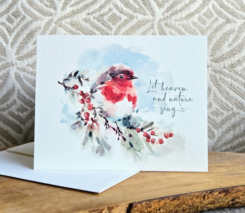 May include: A watercolor greeting card featuring a red-breasted bird perched on a branch with red berries. The card has the text "Let heaven and nature sing" in elegant script, perfect for the holiday season.