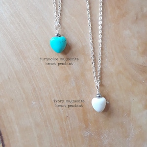 May include: Two heart-shaped magnesite pendants on silver chains. One is turquoise, the other ivory. The text reads "turquoise magnesite heart pendant" and "ivory magnesite heart pendant". The pendants hang against a light brown wooden surface.