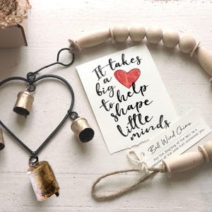 May include: A heart-shaped wind chime with four gold bells and a wooden bead garland. The chime is hanging from a black metal frame. A card with the text "It takes a big heart to help shape little minds" is in the background.