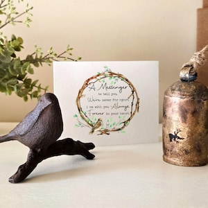 May include: A small, rustic, metal bird figurine perched on a branch. A card with the text "A Messenger to tell you We're never for apart I am with you Always forever in your heart" is in the background. A small, antique-looking bell with a reindeer design hangs from a twine string.