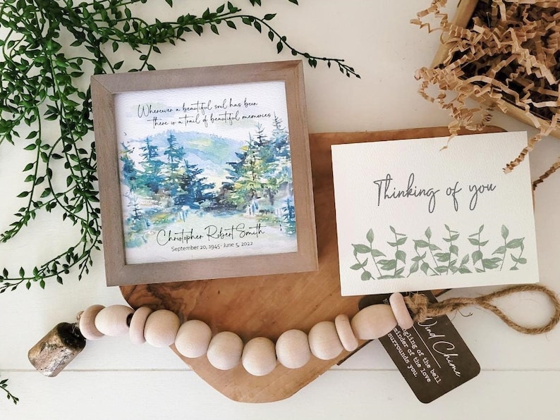 May include: A wooden framed print with a watercolour painting of a forest scene. The print has the text "Wherever a beautiful soul has been, there is a trail of beautiful memories." and "Christopher Robert Smith September 20, 1945 - June 5, 2023". A white card with the text "Thinking of you" and a green floral design is next to the print. A wooden bead garland with a small bell is on a wooden chopping board.