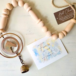 May include: A wooden bell chime with a horse head silhouette inside a copper-colored ring. The chime includes a small bell and a wooden beaded strand. The card reads "Forever in our Hearts" and "Bell Chime".