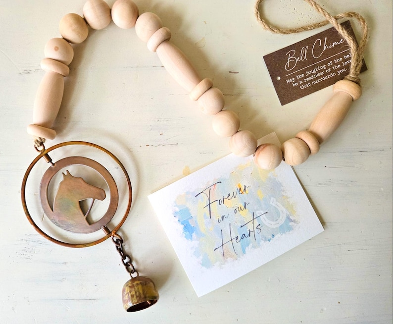 May include: A wooden bell chime with a horse head silhouette inside a copper-colored ring. The chime includes a small bell and a wooden beaded strand. The card reads "Forever in our Hearts" and "Bell Chime".