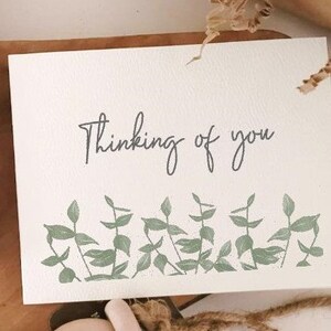 May include: A white card with the text "Thinking of you" in black script. The card is decorated with a border of green leaves.