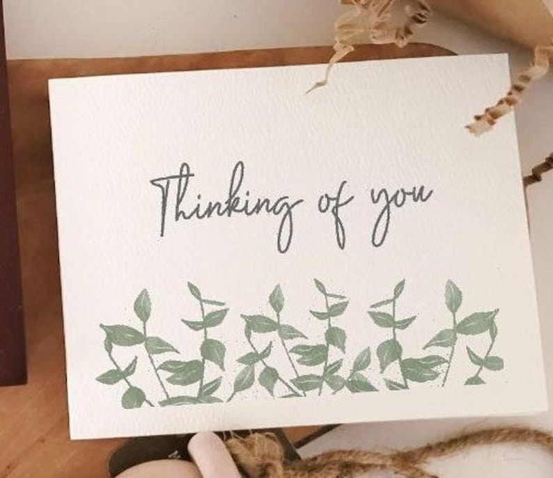 May include: A white card with the text "Thinking of you" in black script. The card is decorated with a border of green leaves.