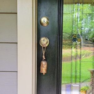 May include: A weathered brass bell hangs from a door handle. The bell is suspended by a natural twine cord. The door has a black finish and a brass doorknob. The glass door panel reflects a green outdoor scene.