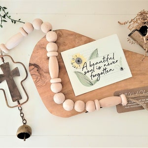 May include: A wooden bead garland with a cross charm and a bell hangs from a wooden cutting board. A card with the text "A beautiful soul is never forgotten" and a sunflower design is on the board. The garland is tied with twine.