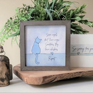 May include: A framed print featuring a blue cat illustration and the text "Some angels don't have wings. Sometimes they have whiskers." A sympathy card reads "So sorry for your loss." A rustic bell and greenery are also present.