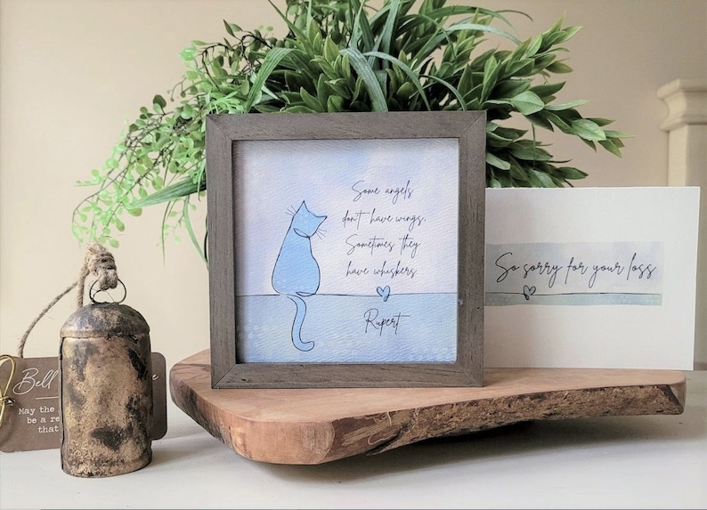 May include: A framed print featuring a blue cat illustration and the text "Some angels don't have wings. Sometimes they have whiskers." A sympathy card reads "So sorry for your loss." A rustic bell and greenery are also present.