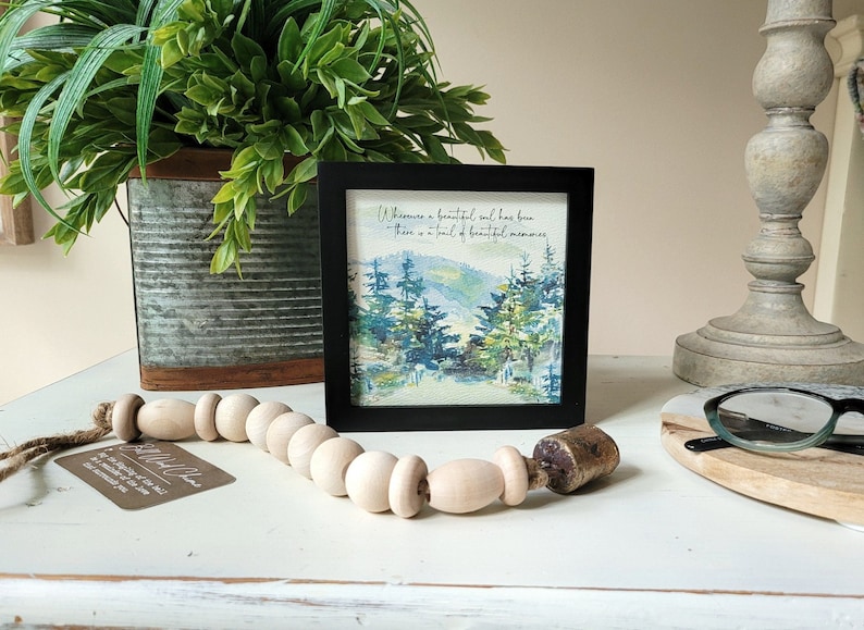 May include: A framed watercolour painting of a forest scene with the quote "Where there is a beautiful soul, there is a trail of beautiful memories." The frame is black and the painting is on a white background.