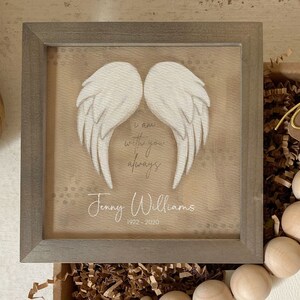 May include: A framed wooden sign with a white painted angel wing design. The sign has a beige background with brown speckles and the text "I am with you always" and "Jenny Williams 1922-2020".