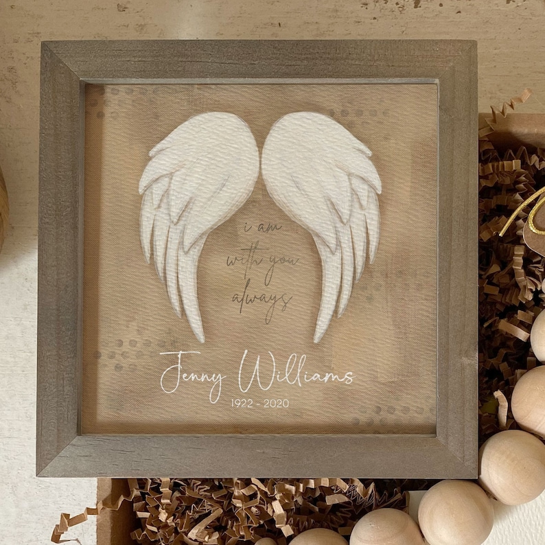 May include: A framed wooden sign with a white painted angel wing design. The sign has a beige background with brown speckles and the text "I am with you always" and "Jenny Williams 1922-2020".