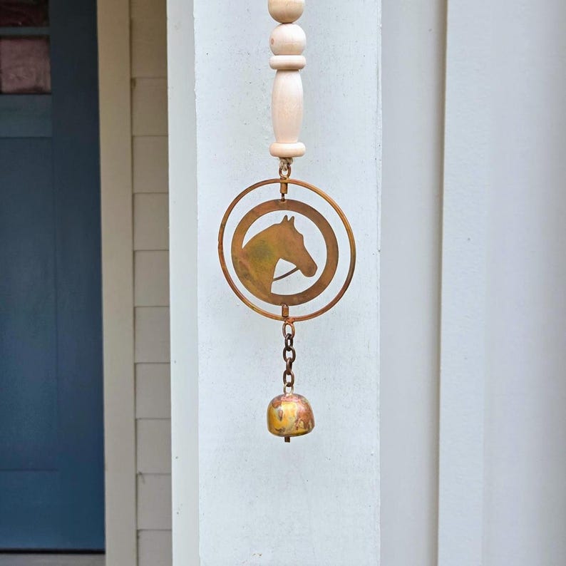 May include: A decorative wind chime featuring a horse head silhouette within a circular frame. The chime is made of copper-colored metal and hangs from a wooden top with a small bell at the bottom. The design is suitable for outdoor decor.