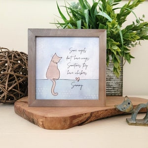 May include: A framed print with a light brown frame, featuring a light pink cat illustration and the text "Some angels don't have wings. Sometimes they have whiskers." The print is displayed on a wooden surface.