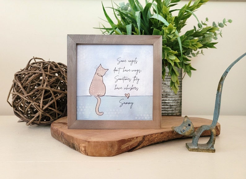 May include: A framed print with a light brown frame, featuring a light pink cat illustration and the text "Some angels don't have wings. Sometimes they have whiskers." The print is displayed on a wooden surface.