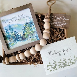 May include: A gift box filled with brown packing paper, a wooden bead garland with a bell, a framed watercolour painting of a forest scene, and a card that reads "Thinking of you" with a green leaf design.