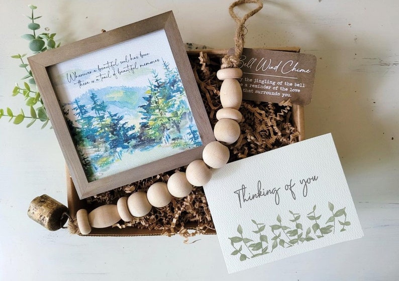 May include: A gift box filled with brown packing paper, a wooden bead garland with a bell, a framed watercolour painting of a forest scene, and a card that reads "Thinking of you" with a green leaf design.