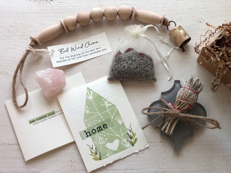 May include: A home blessing kit with a sage smudge stick, a pink rose quartz crystal, a small bag of lavender, a wooden bell wind chime, and a card that says "Home Blessing Guide" with joy, laughter, abundance, and more.