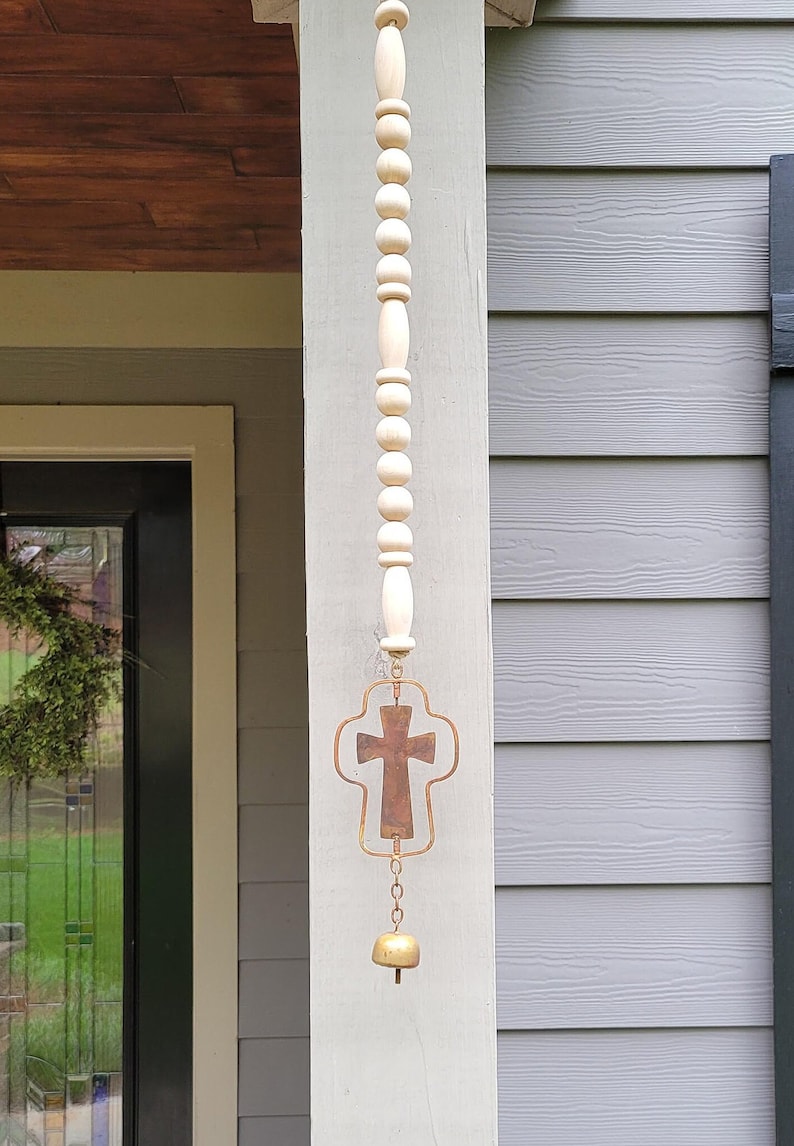 K&ouml;nnte beinhalten: Dekorative Windglocke mit einer Holzkette aus Perlen, einem Metallkreuz und einer kleinen Messingglocke. Das Kreuz hat eine rustikale Bronzeoberfl&auml;che. Die Holzperlen sind hell.