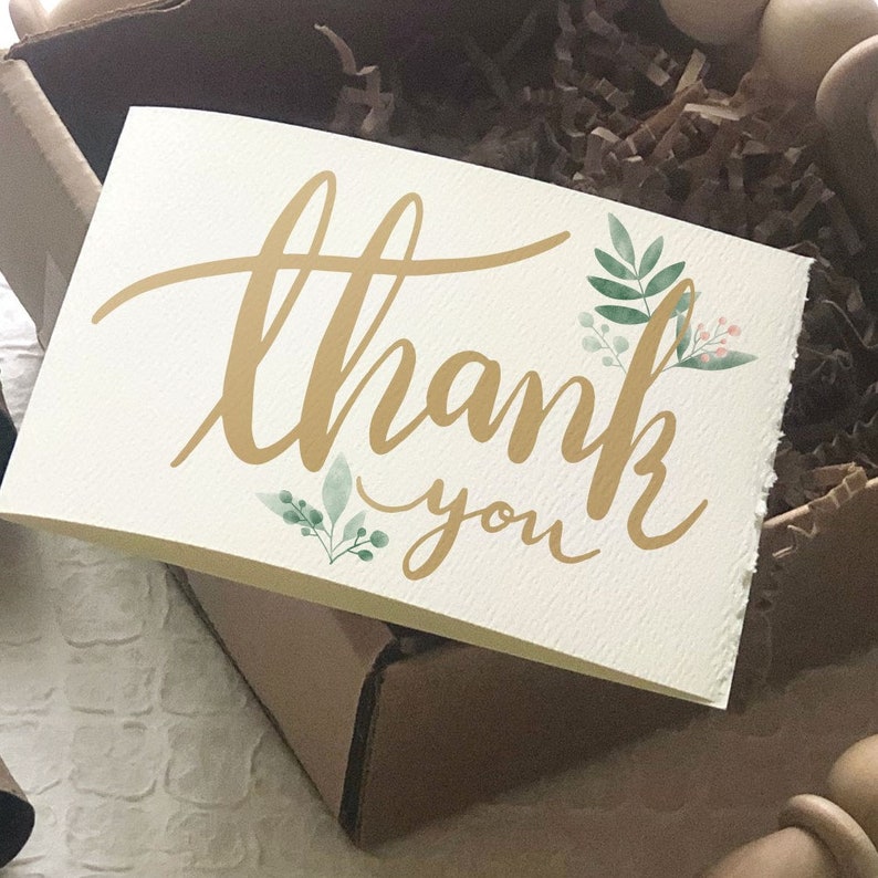 K&ouml;nnte beinhalten: Eine cremefarbene Dankeskarte mit goldener Kalligraphie und Aquarell-Bl&uuml;tenakzenten. Die Karte befindet sich in einer braunen Pappschachtel mit braunem Papierf&uuml;llmaterial. Die Kartenr&auml;nder sind rau.