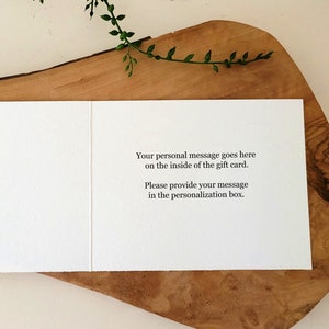 May include: An open white greeting card with text: "Your personal message goes here on the inside of the gift card. Please provide your message in the personalization box." The card is displayed on a wooden board, with greenery in the background.