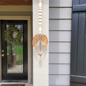 May include: A rustic metal angel wing wind chime with a bell hanging from a chain. The chime is attached to a wooden bead garland.