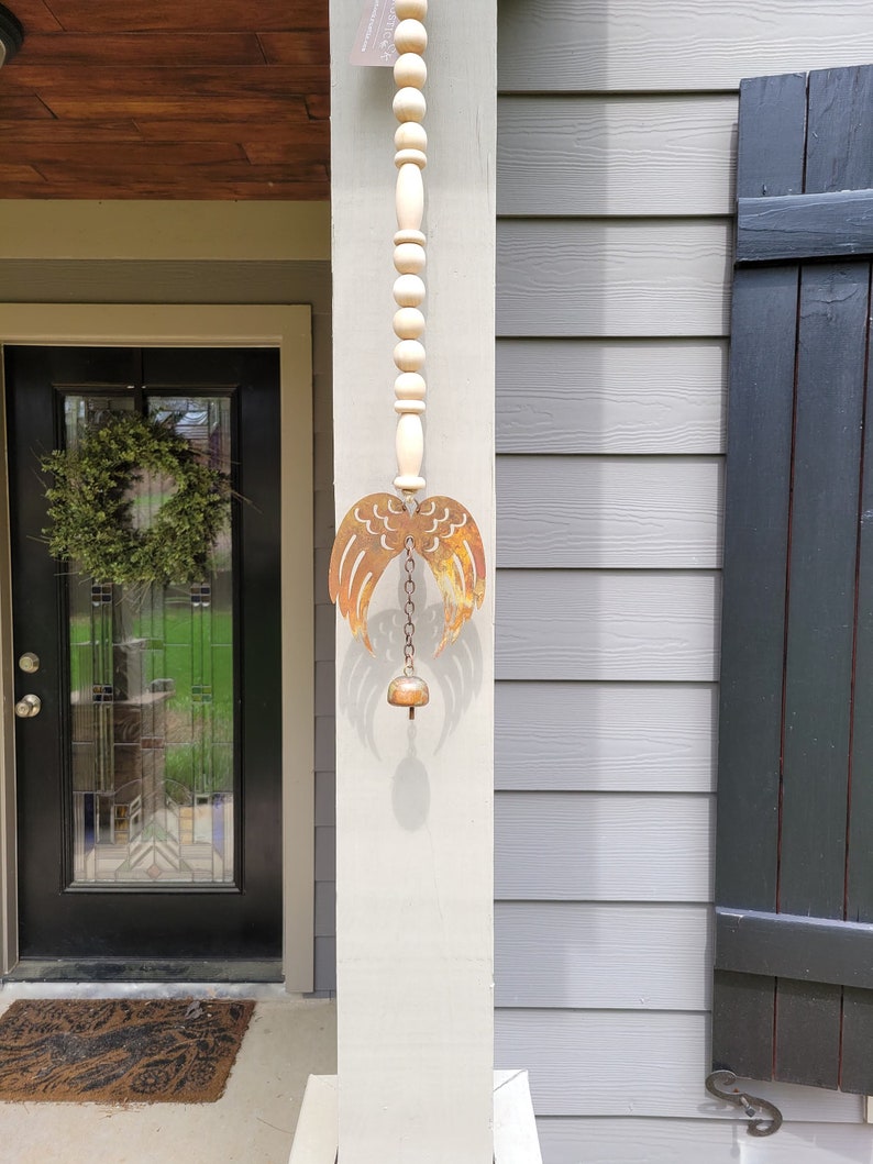 May include: A rustic metal angel wing wind chime with a bell hanging from a chain. The chime is attached to a wooden bead garland.