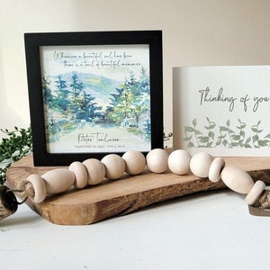 May include: A framed watercolour painting of a forest scene with the quote "Wherever a beautiful soul has been, there is a trail of beautiful memories."  The painting is signed "Peter Tomlinson" and dated "September 29, 1945 - June 5, 2023". A wooden bead garland with a gold bell is on a wooden surface in front of the painting. A card with the text "Thinking of you" is on the right side of the image.