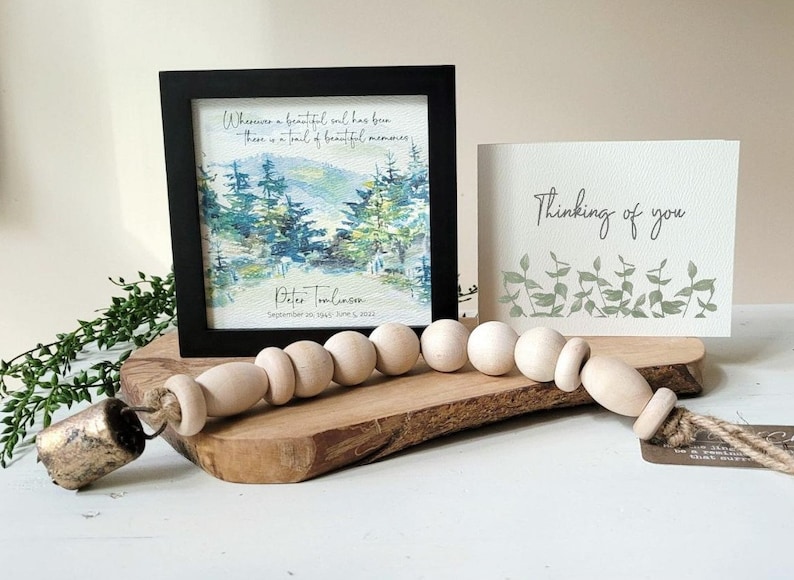 May include: A framed watercolour painting of a forest scene with the quote "Wherever a beautiful soul has been, there is a trail of beautiful memories."  The painting is signed "Peter Tomlinson" and dated "September 29, 1945 - June 5, 2023". A wooden bead garland with a gold bell is on a wooden surface in front of the painting. A card with the text "Thinking of you" is on the right side of the image.